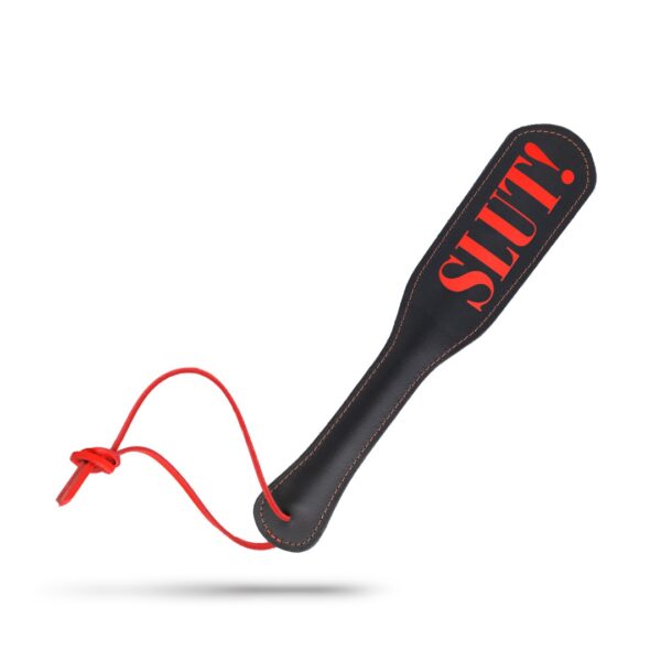 Na obrazie jest skórzana czarna packa z czerwonym napisem napis "SPANK ME" . O"Uderz mnie" l„Daj mi klapsa” .